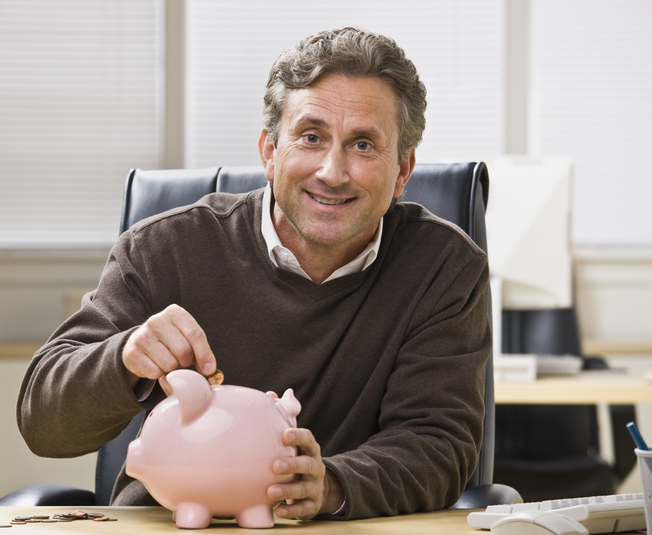 A man is seated at a desk in an office and is putting a coin into a piggy bank.  He is smiling at the camera.  Horizontally framed shot.