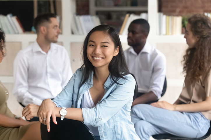 Asian appearance woman psychologist addiction counsellor smile look at camera, on background diverse young people sitting in circle share problems supporting each other concept of rehab center session