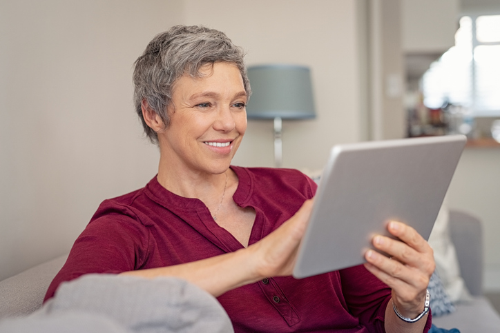 Smiling senior woman looking her digital tablet while sitting on sofa. Portrait of mature happy woman relaxing at home with digital tablet. Happy lady with gray hair browsing on laptop in living room.