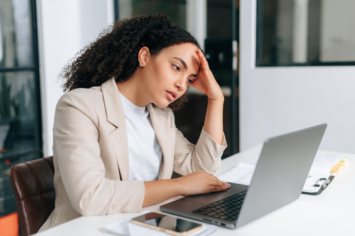 Sad brazilian or hispanic curly haired business woman, corporate financial manager, sits in a modern office, sadly looking at a laptop screen, reading bad news, received bad financial report
