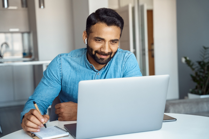 Portrait of young adult indian man wearing earphones looking at laptop screen. Motivated student writing notes during online lesson, watching webinar, learning language online, sitting at work desk